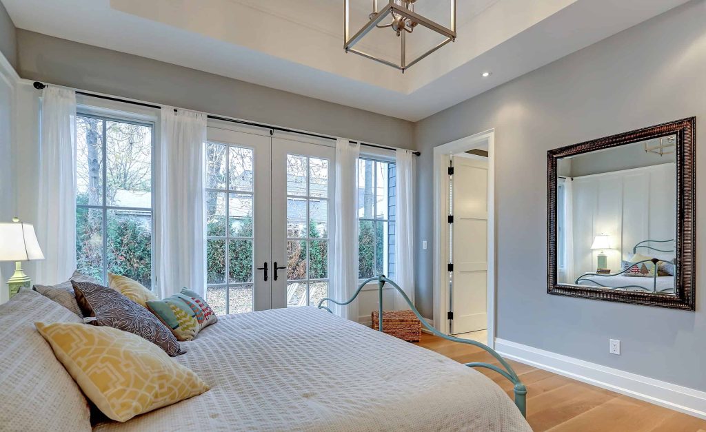 A bedroom in one of the charming Houses For Sale in Oakville, boasting large windows, a double bed with patterned pillows, a wall mirror, and a bedside lamp. The room features light gray walls and a delightful mix of natural and artificial lighting, chatsworth construction