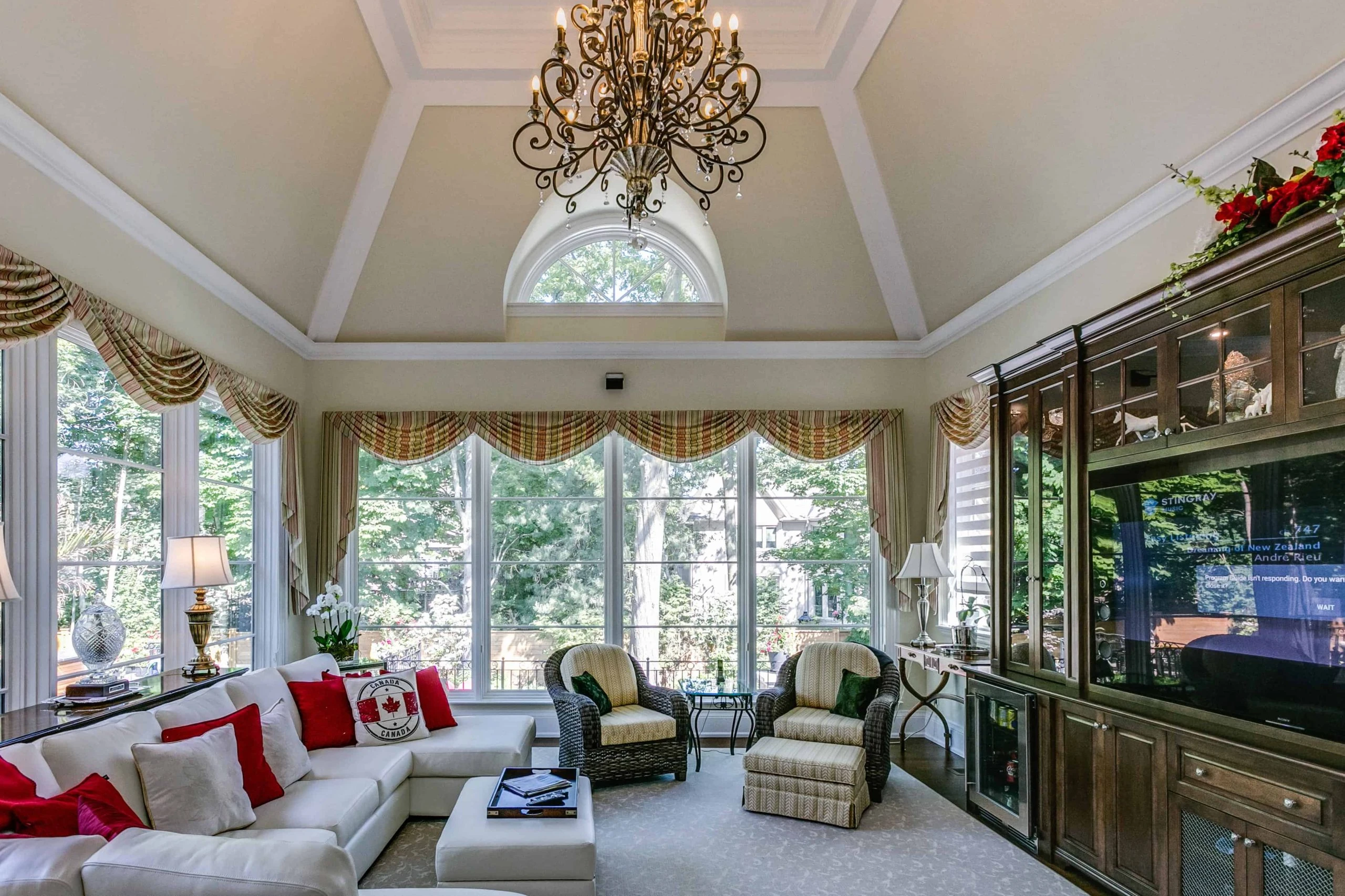 A spacious living room with large windows, a vaulted ceiling featuring a chandelier, and furnished with white sofas, red pillows, a glass coffee table, and a large dark wood entertainment unit is the hallmark of luxury custom home builders.