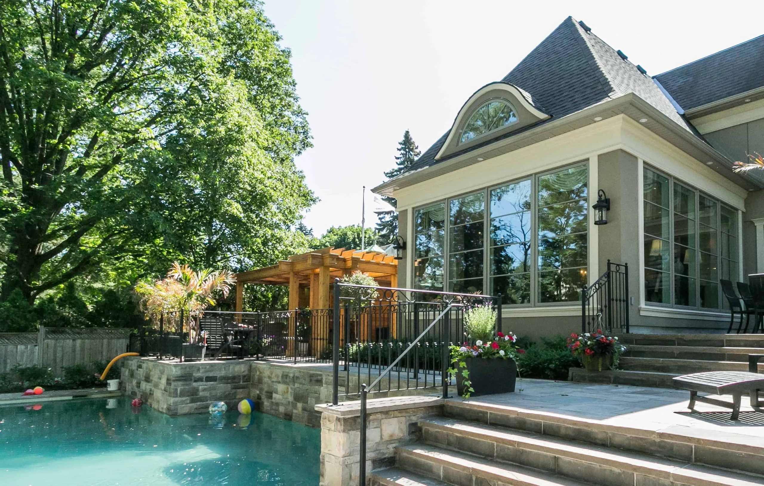 A residential backyard in Oakville luxury real estate featuring a swimming pool, patio with stone steps, potted plants, and a wooden pergola near a house with large windows.