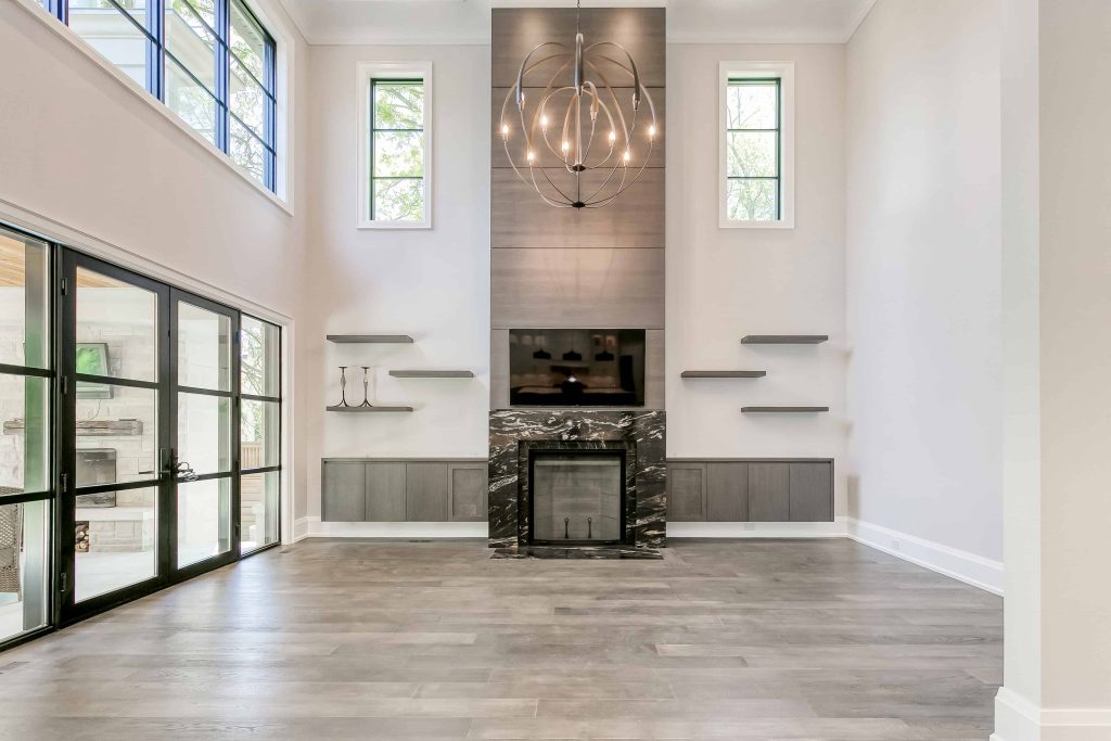 Spacious modern living room with high ceilings, a large chandelier, a fireplace with a wall-mounted TV, built-in shelves, large windows, and glass doors providing an abundance of natural light—a perfect highlight for anyone searching for houses for sale in Oakville, custom home builder mississauga