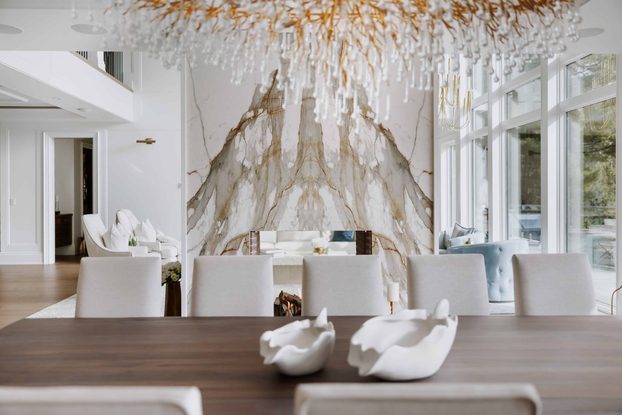 A modern dining area in custom homes in Oakville features a large wooden table, white cushioned chairs, abstract white centerpieces, a striking marble wall feature, and a grand chandelier with a nature-inspired design.