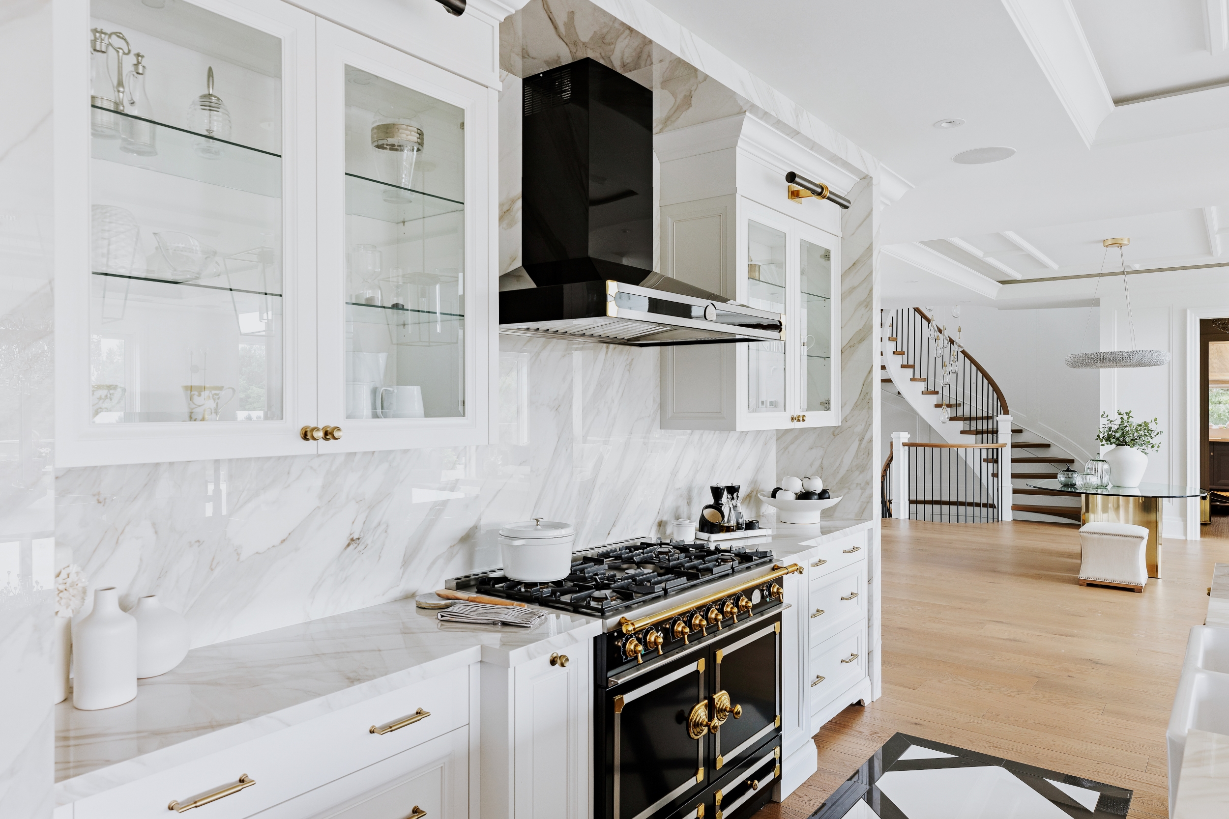 Explore the modern kitchen at East Caledon Estate, featuring sleek white cabinets, a striking black and gold stove, and an elegant marble backsplash. In the background, stylish stairs lead to a sophisticated dining area.