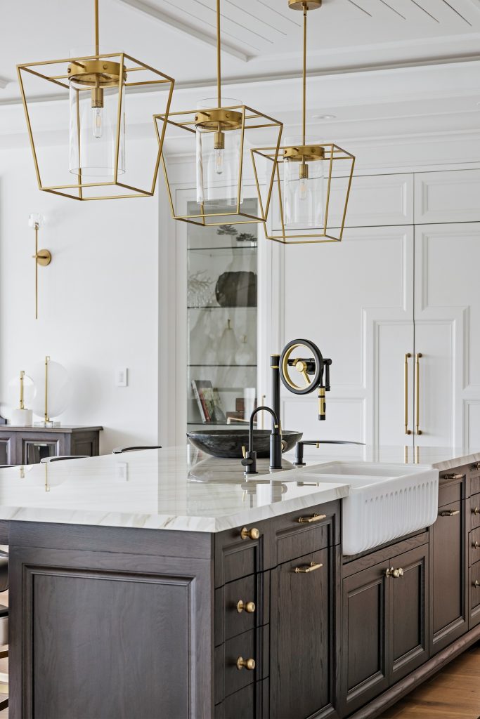 A modern Kitchen Burlington features a large island with a marble countertop, dark cabinetry, gold hardware, a farmhouse sink, and three geometric gold pendant lights above, Kitchen Burlington