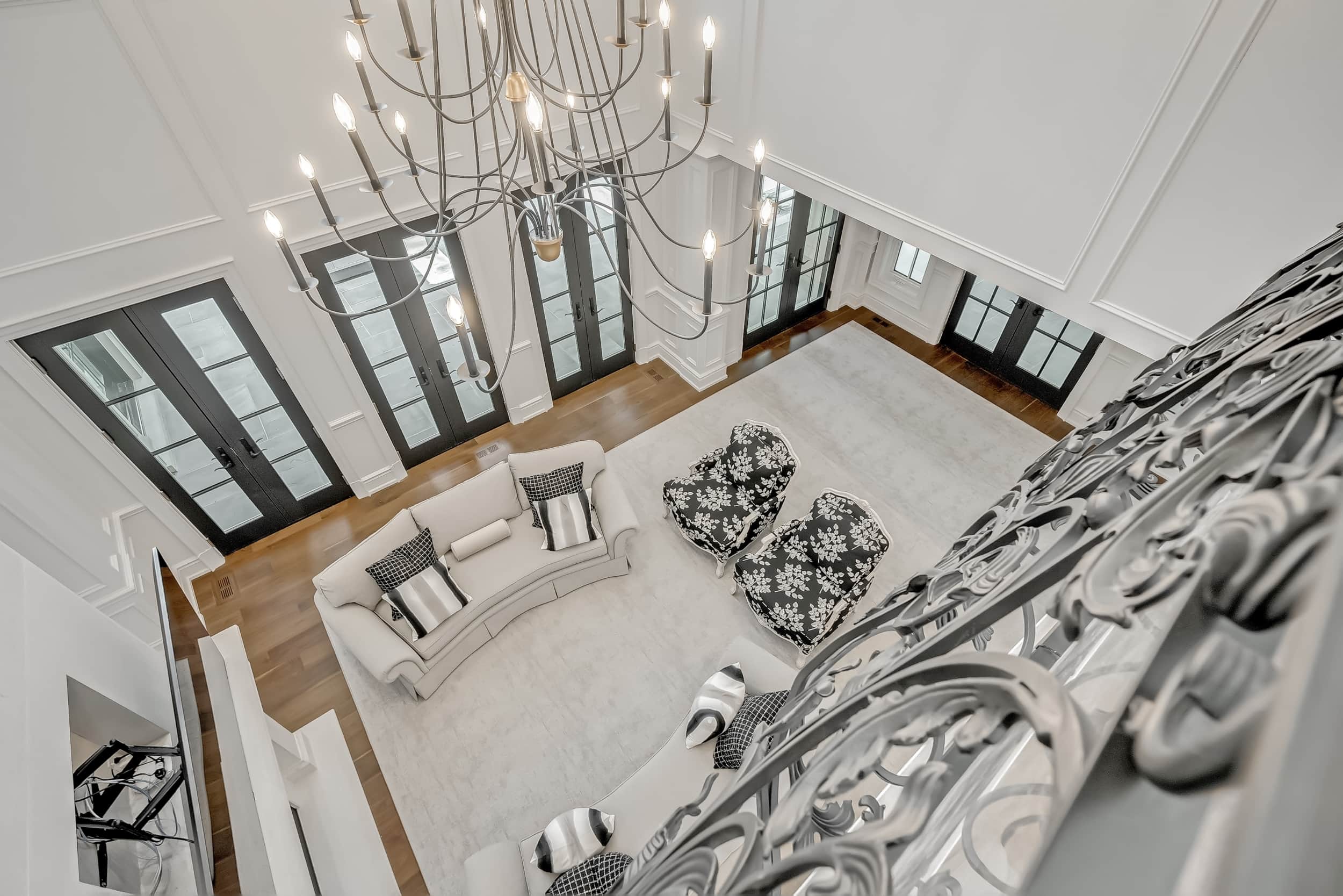 Aerial view of a spacious living room in a custom luxury home in Oakville, featuring white furniture, black patterned armchairs, a large chandelier, hardwood floors, and tall black-framed windows.