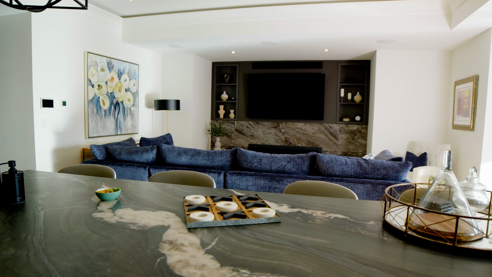 A custom home in Oakville features a modern living room with a large blue sectional couch, a TV mounted on a wall with built-in shelves, a marble countertop, a decorative painting, and various elegant items.