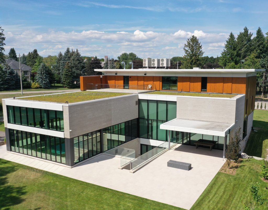 Modern two-story house with large glass windows, flat roofs, and a mix of white and wooden exterior panels, built using eco-friendly materials. It is surrounded by a well-maintained lawn and trees.