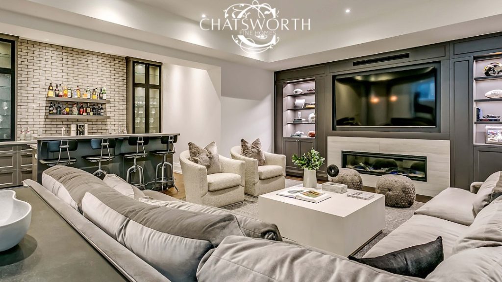 Modern living room featuring a large sectional sofa, two armchairs, a fireplace, a wall-mounted TV, and a bar area with stools. Showcasing "Chatsworth Fine Homes," known for sustainable custom luxury homes using eco-friendly materials.