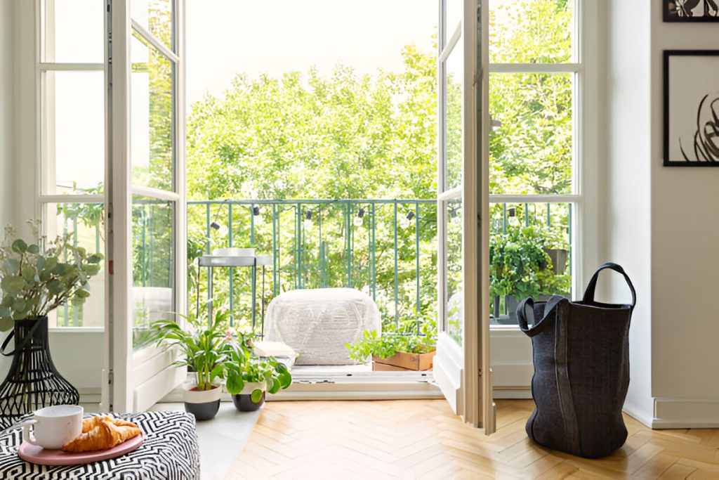 A bright room with an open balcony door, various plants, a black bag on the floor, and a blanket-covered chair outside. A table crafted from eco-friendly materials with a cup and a plate of food is on the left. This space exemplifies the charm of sustainable custom luxury homes.