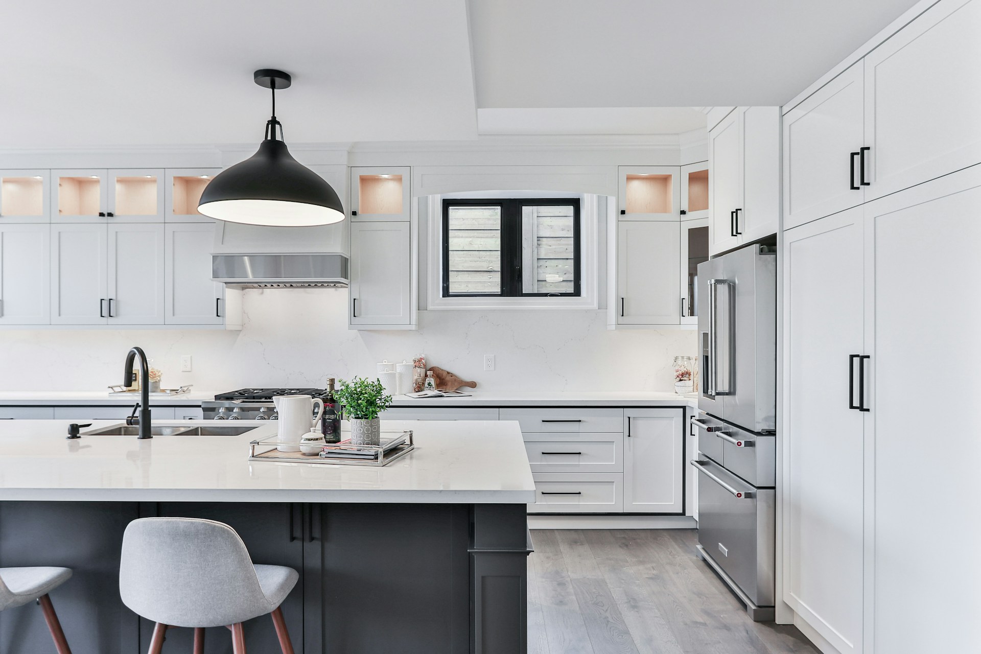 Modern kitchen in Oakville featuring white cabinets, stainless steel appliances, a large island with a white countertop, and a black pendant light. A small tray with decor sits on the island, and a window is above the sink, Kitchen Oakville