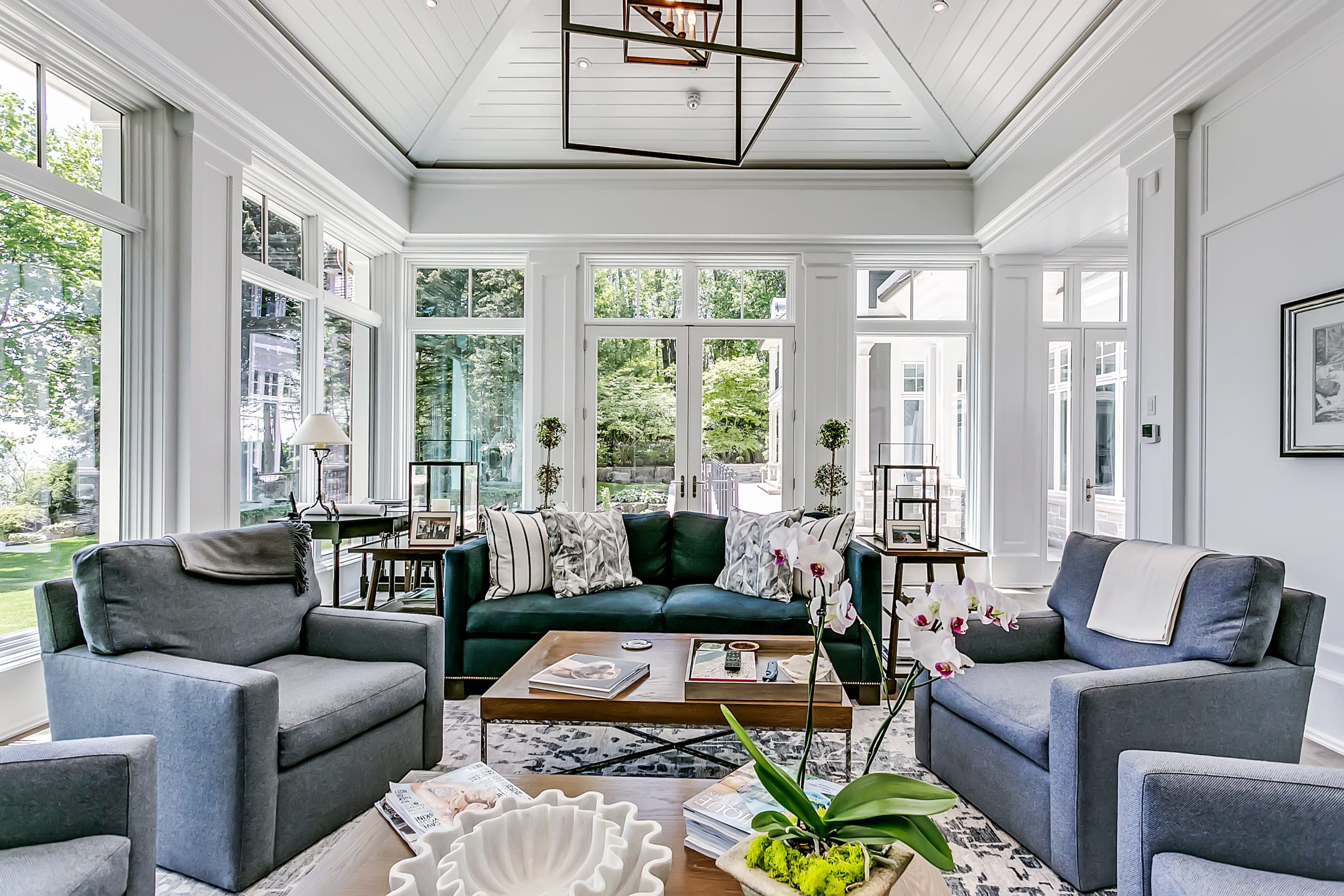 A modern, light-filled living room with large windows, gray armchairs, a green sofa, a wooden coffee table adorned with decorative items, and potted plants. Discover this inviting space among our stunning homes for sale.