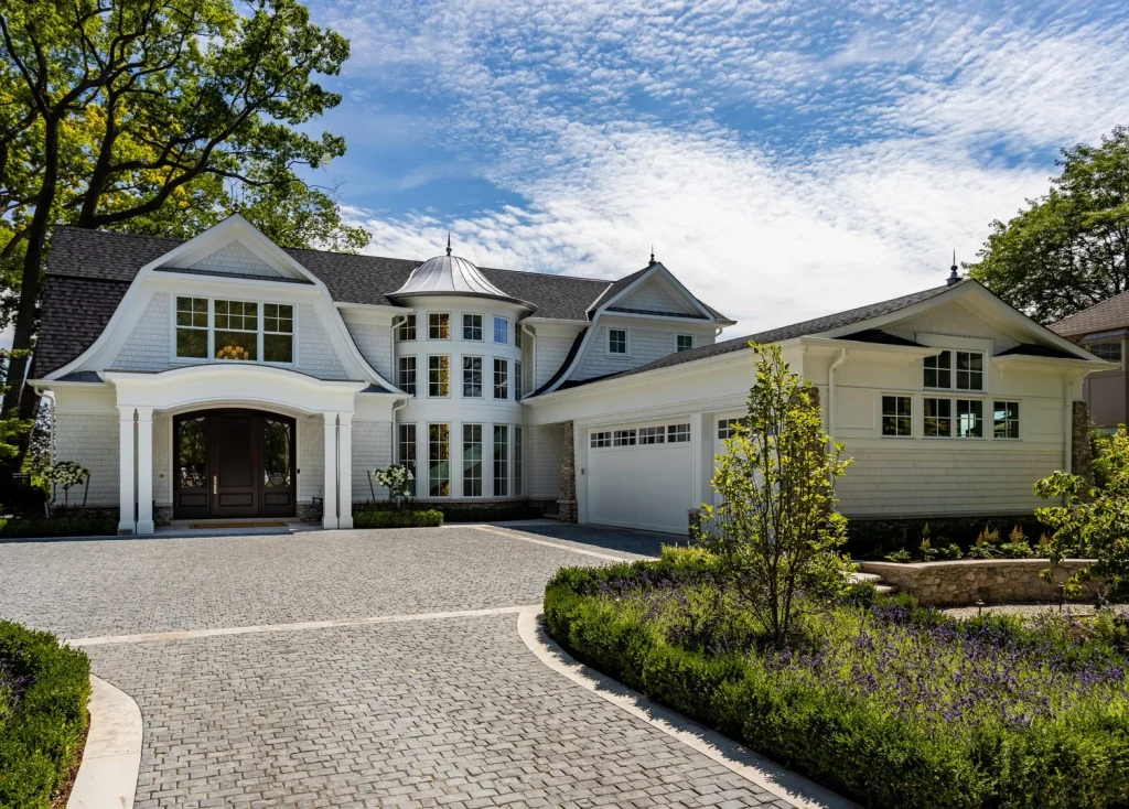 A large white house with multiple gables, a circular driveway, and attached garage is surrounded by manicured landscaping and trees under a partly cloudy sky—a perfect example of luxury homes building at its finest, Real Estate Oakville