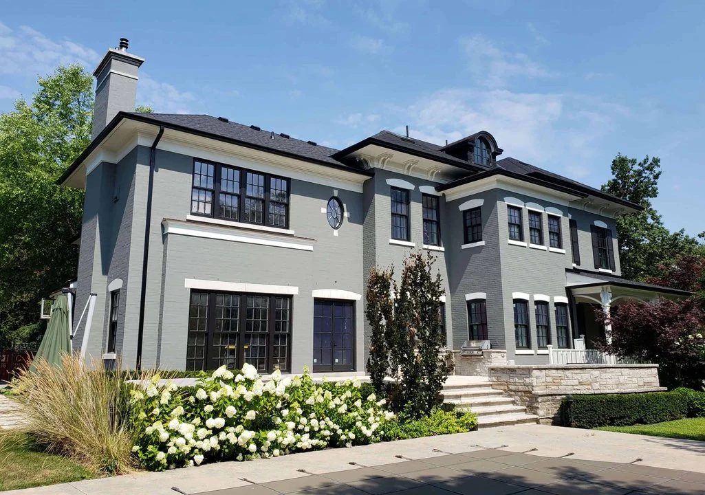 A two-story grey house with black shutters, white trim, and a small front porch, surrounded by green shrubs and plants under a clear blue sky exemplifies our expertise in building luxury homes.