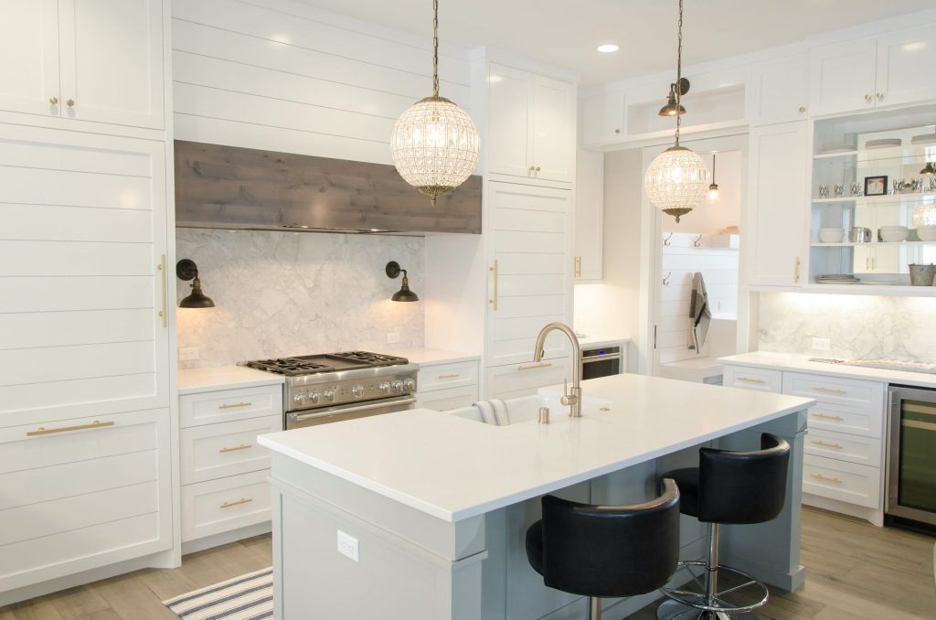 A modern kitchen with white cabinets, a marble backsplash, two pendant lights, a central island with a sink, black bar stools, stainless steel appliances including a stove and wine cooler—perfect for those looking for kitchen design ideas.