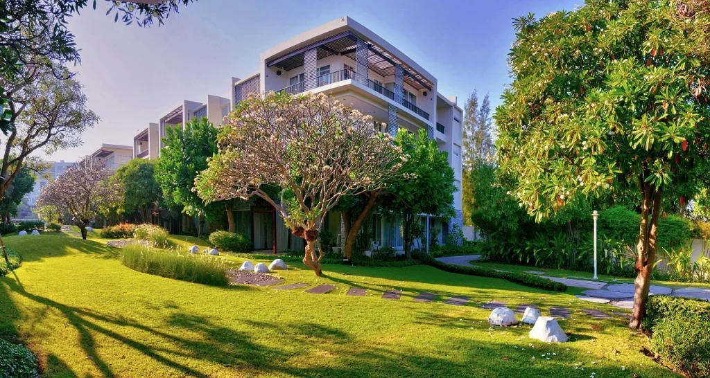 A modern three-story building with balconies, serving as the ideal location for your custom home, is surrounded by a well-manicured lawn, trees, and landscaped pathways on a sunny day.