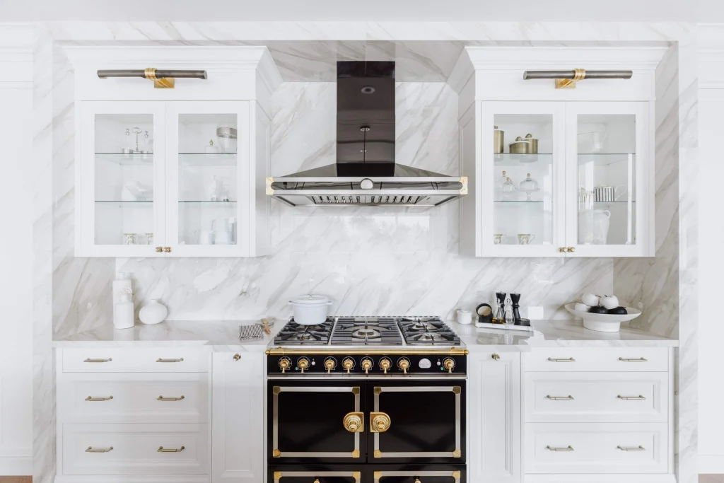 A modern kitchen with a black and gold stove, white marble backsplash, glass-front cabinets, and white countertops. For those seeking Kitchen Design Ideas, cookware and decorative items are visible inside the cabinets and on the countertop.