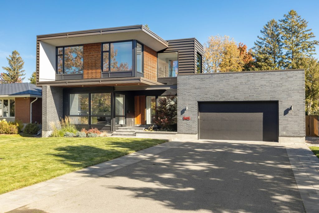 A modern two-story house with large windows, a mix of brick and wood exterior, and a double garage, exemplifies the art of building luxury homes. Situated on a sunny day with a neat lawn and driveway, it boasts an exquisite backdrop of trees.