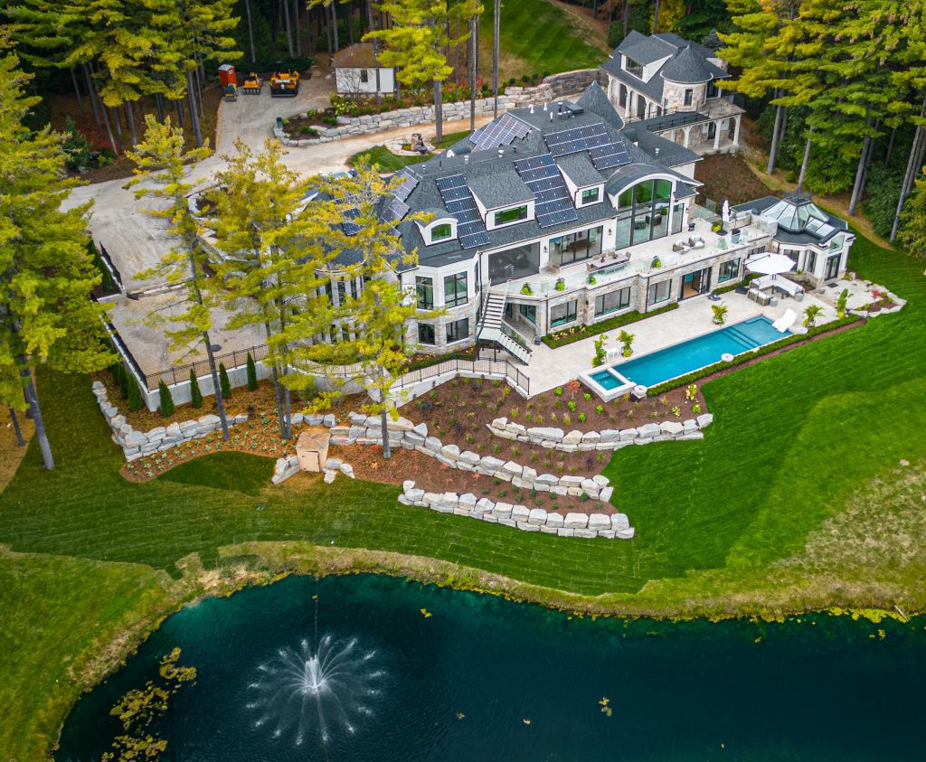 Aerial view of a large, modern mansion with a swimming pool, landscaped garden with stone terraces, and a small pond with a fountain, surrounded by trees—an ideal location for your custom home.