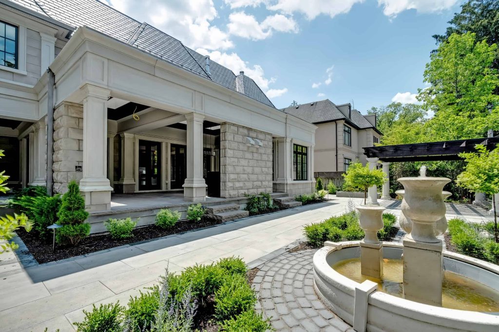 A luxurious two-story house with a stone facade featuring a covered entrance, landscaped garden, and a fountain in the foreground. This prime example of building luxury homes is surrounded by greenery and set against a vivid blue sky.