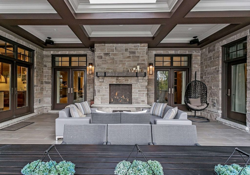 Experience outdoor luxury living on this covered patio featuring a stone fireplace, gray sofas, decorative pillows, a hanging chair, large windows, and a wooden table with potted plants in the foreground.