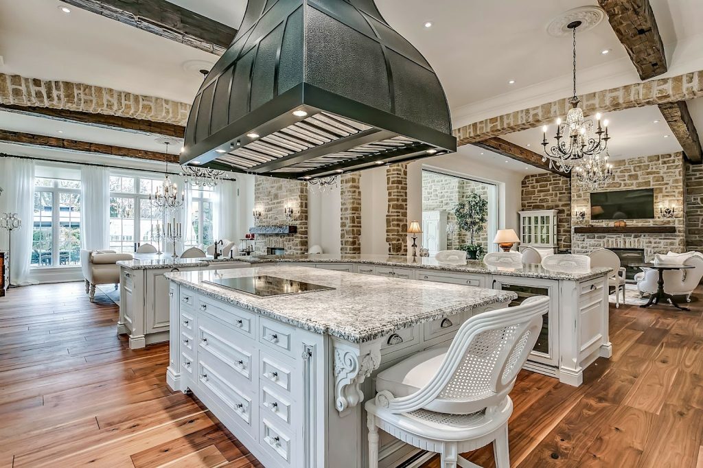 Spacious kitchen with marble countertops, large island, ornate white cabinetry, wood floors, exposed brick walls, chandeliers, and a large black range hood—perfect for those who appreciate indoor elegance and outdoor luxury living.
