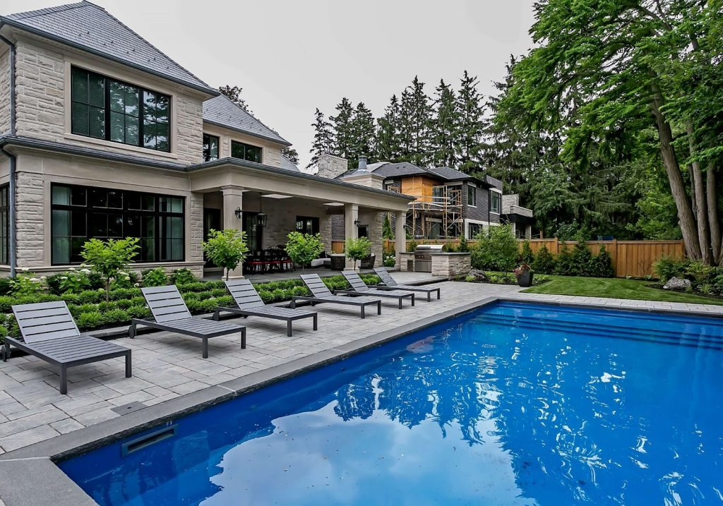 Modern house with large patio, six lounge chairs, and an in-ground swimming pool surrounded by trees and landscaped greenery—perfect for embracing outdoor luxury living.