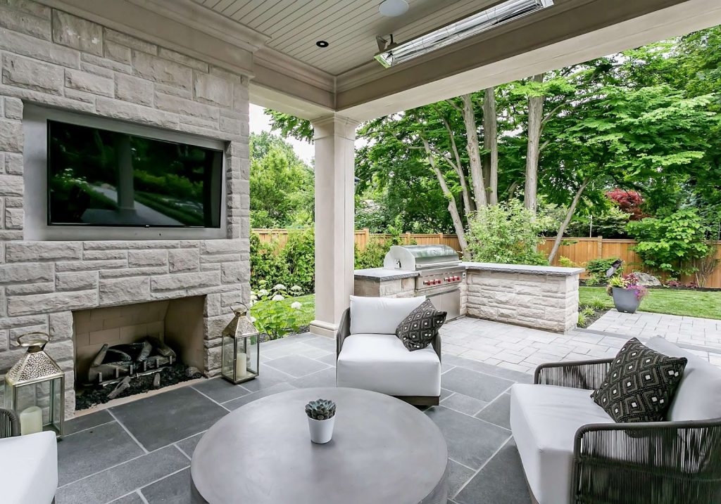 Covered patio with stone fireplace, mounted TV, two chairs, round coffee table, outdoor kitchen, and garden with trees and shrubs in the backgroundโperfect for embracing the latest Outdoor Living Trends.