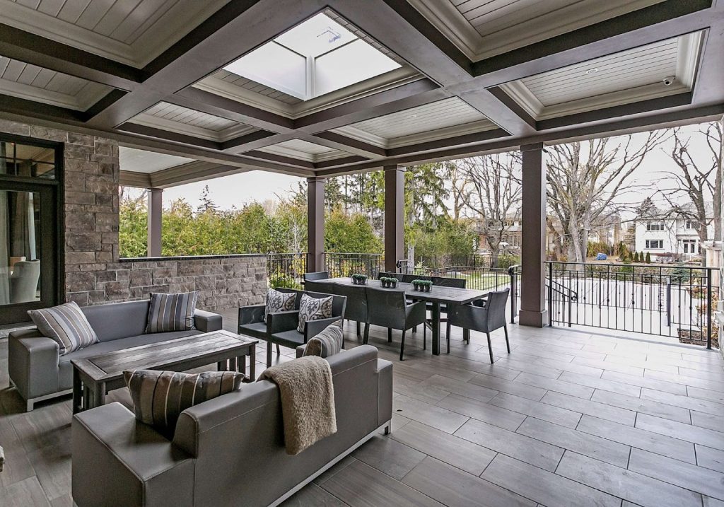 A covered outdoor patio with modern seating, a dining table, and neutral-toned furniture reflects the latest Outdoor Living Trends, all while offering views of trees and neighboring houses.