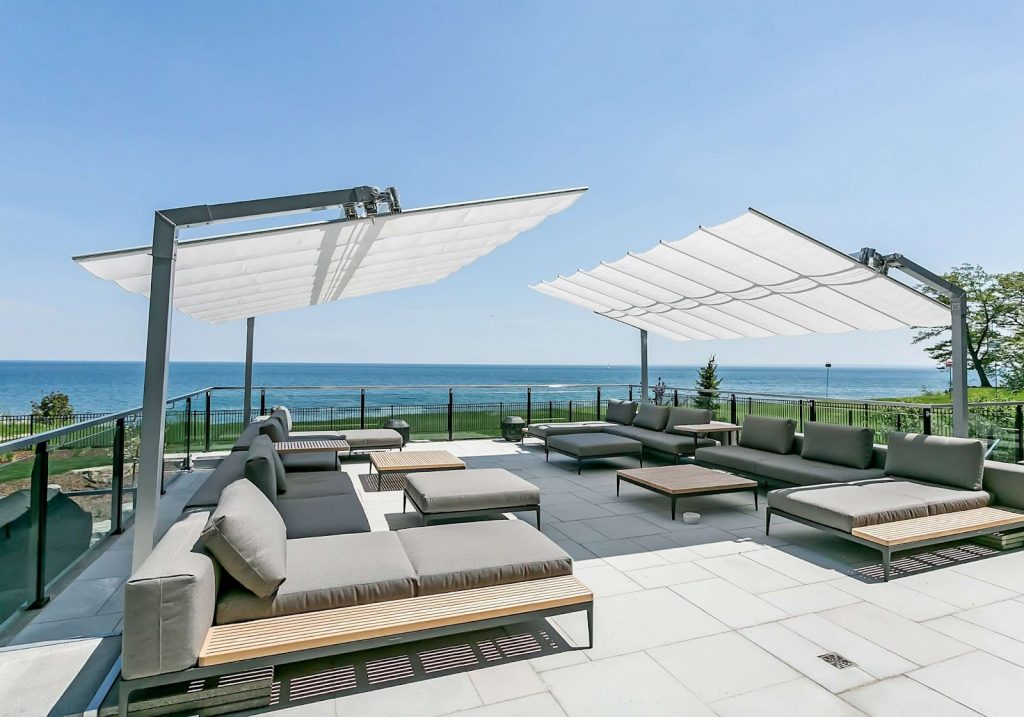 Modern outdoor patio with gray cushioned seating, wooden tables, and two large white retractable canopies showcases Outdoor Living Trends as it overlooks a body of water on a clear, sunny day.