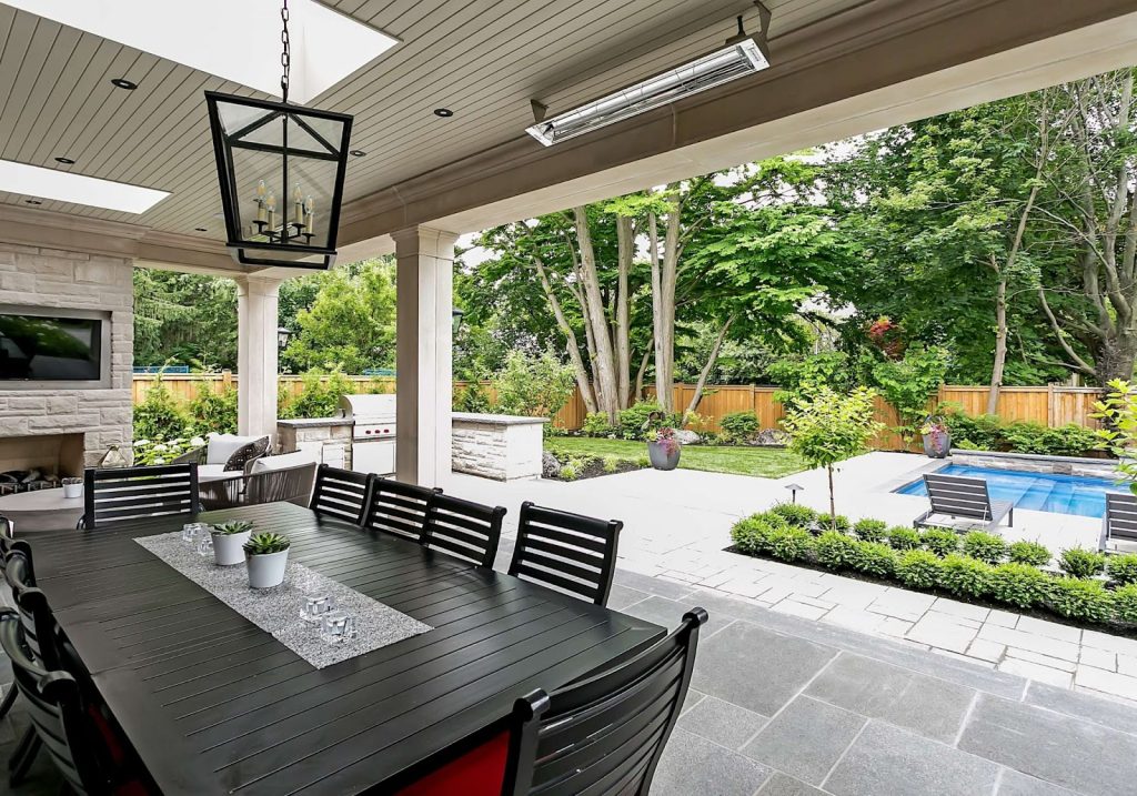 Covered patio with black dining table and chairs overlooks a landscaped backyard featuring a pool, grill area, lush greenery, and wood fence—embracing the latest Outdoor Living Trends.